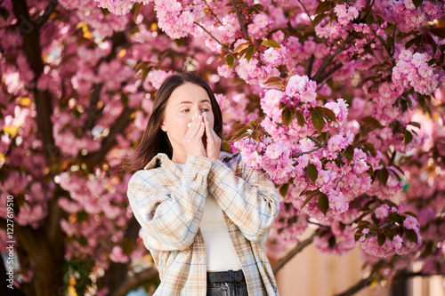 Woman allergic suffering from seasonal allergy at spring in blossoming garden at springtime. Woman sneezing and blowing nose using nasal handkerchief in front of blooming tree. Spring allergy concept. photo