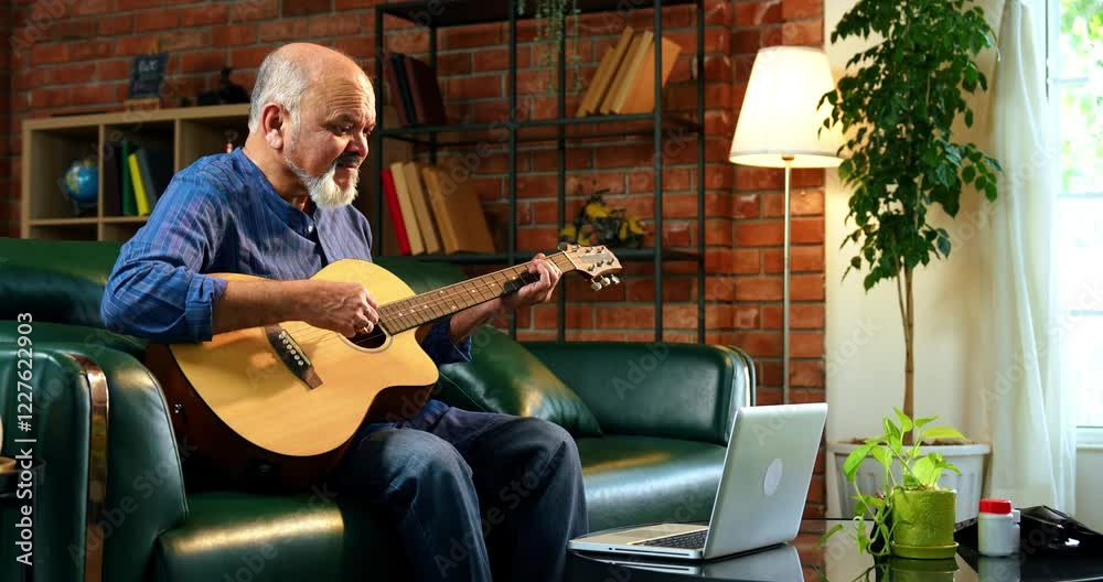 Indian senior retired man with a beard wearing kurta, sitting on a sofa in a modern living room, learning to play acoustic guitar from an online tutorial on his laptop, enjoying music practice