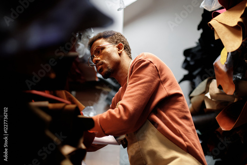 Man organizing leather materials in well-lit workshop, showing keen focus. Leather pieces and supplies are visible in neatly arranged shelves in background photo