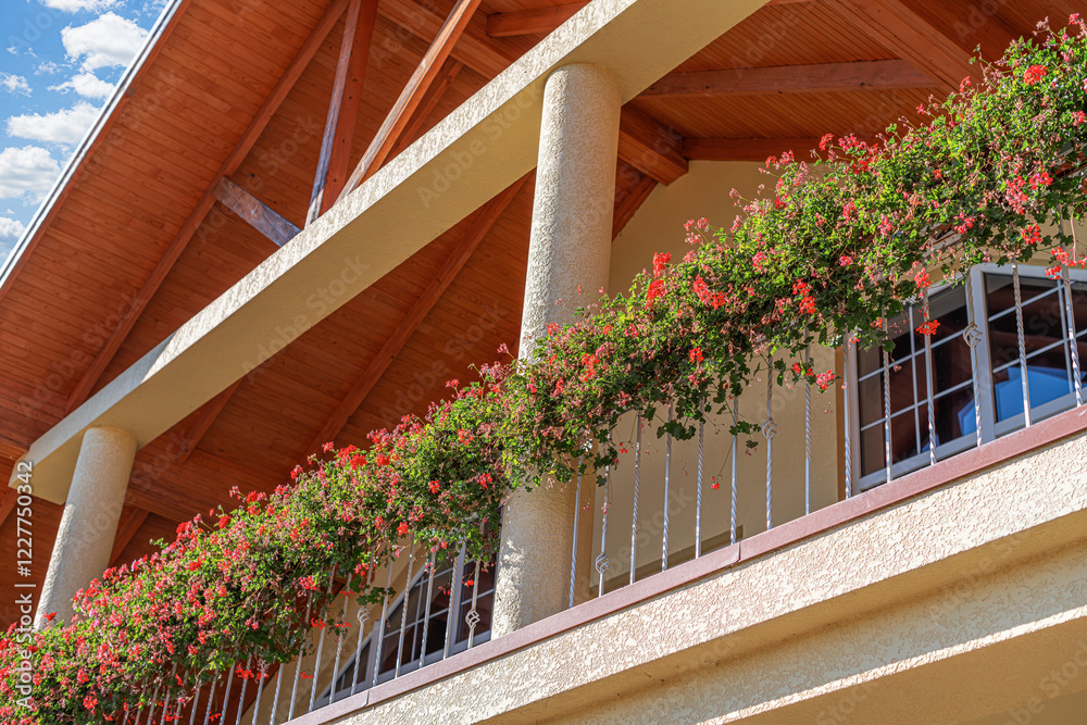 A beautifully designed balcony with vibrant red flowers cascading over the railing. The wooden roof and textured columns add a rustic charm to the architecture under a bright, sunny sky.