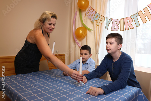 A woman with her son and nephew decorate a room and a festive table for a birthday celebration. Soft lighting.