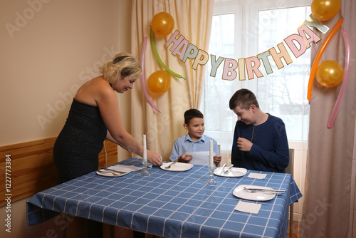 A woman with her son and nephew decorate a room and a festive table for a birthday celebration. Soft lighting.
