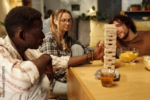Side view of youthful guy looking at tower built up on table while playing tabletop game with friends