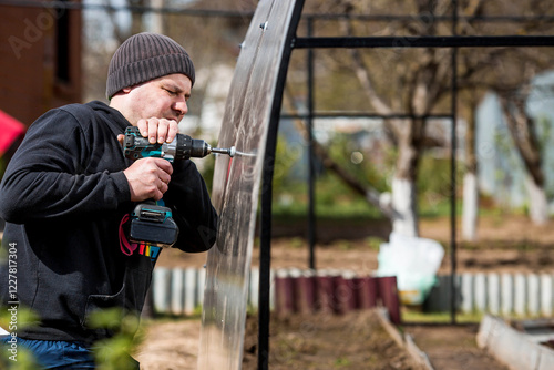 A man holds a battery-powered screwdriver and assembles a new greenhouse made of polyethylene, polycarbonate. Macro photography. Side view. Preparing for the spring garden season.