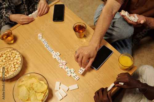 High angle of hands of three young intercultural friends playing domino by wooden table at leisure
