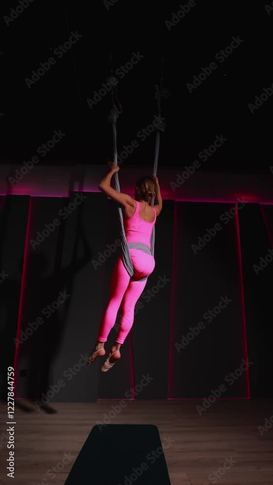 Aerial yoga enthusiast skillfully performs graceful maneuvers on silks, highlighting balance and body control in a dimly lit studio with pink accents Aerial yoga enthusiast skillfully performs graceful maneuvers on silks, highlighting balance and body control in a dimly lit studio with pink accents