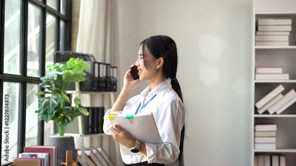 Businesswoman on the Phone: A young, professional Asian businesswoman smiles warmly as she speaks on her mobile phone, holding documents in her other hand. Businesswoman on the Phone: A young, professional Asian businesswoman smiles warmly as she speaks on her mobile phone, holding documents in her other hand.