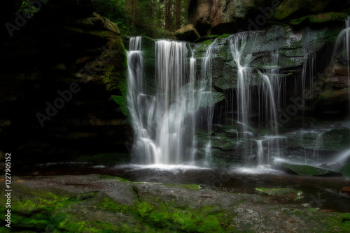 A serene waterfall flows gracefully over moss-covered rocks, surrounded by lush greenery.