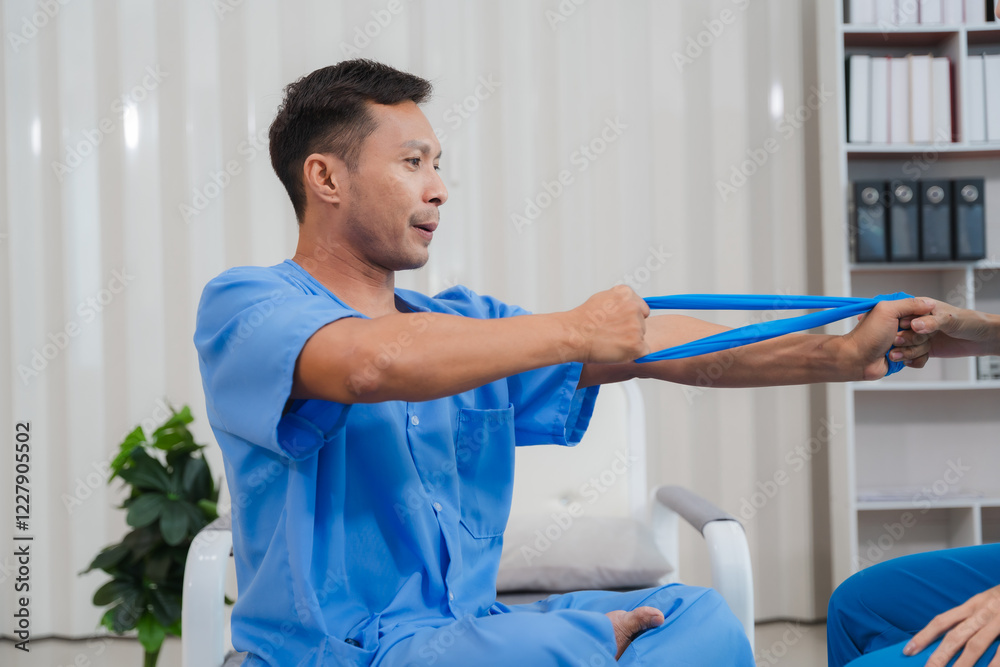 Physical Therapy Resistance Band Exercise: A male patient wearing a blue medical gown participates in physical therapy, using a blue resistance band to stretch and strengthen his arm. Physical Therapy Resistance Band Exercise: A male patient wearing a blue medical gown participates in physical therapy, using a blue resistance band to stretch and strengthen his arm.