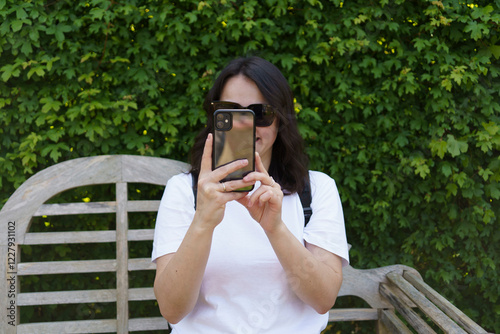 Beautiful smiling dark-haired girl in glasses with a backpack and in a white T-shirt takes a photo sitting on a wooden bench in a summer park. Concept of capturing a moment of life photo