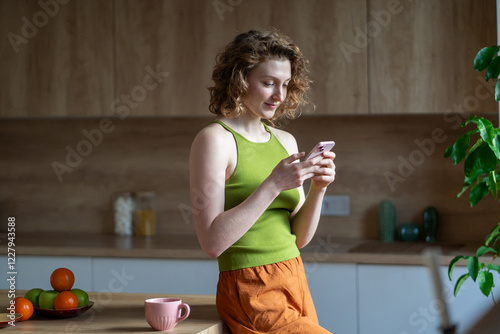 Calm smiling woman enjoying slow morning in kitchen, scrolling social media on smartphone. Relaxed pleased European young female using phone reading news or chatting with friends standing at cozy home