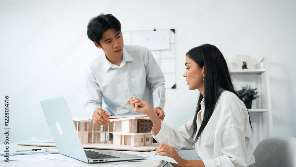 Professional male asian architect using ruler to measure house model length while young beautiful caucasian colleague using laptop to analyzed data on meeting table with house model. Immaculate. Professional male asian architect using ruler to measure house model length while young beautiful caucasian colleague using laptop to analyzed data on meeting table with house model. Immaculate.