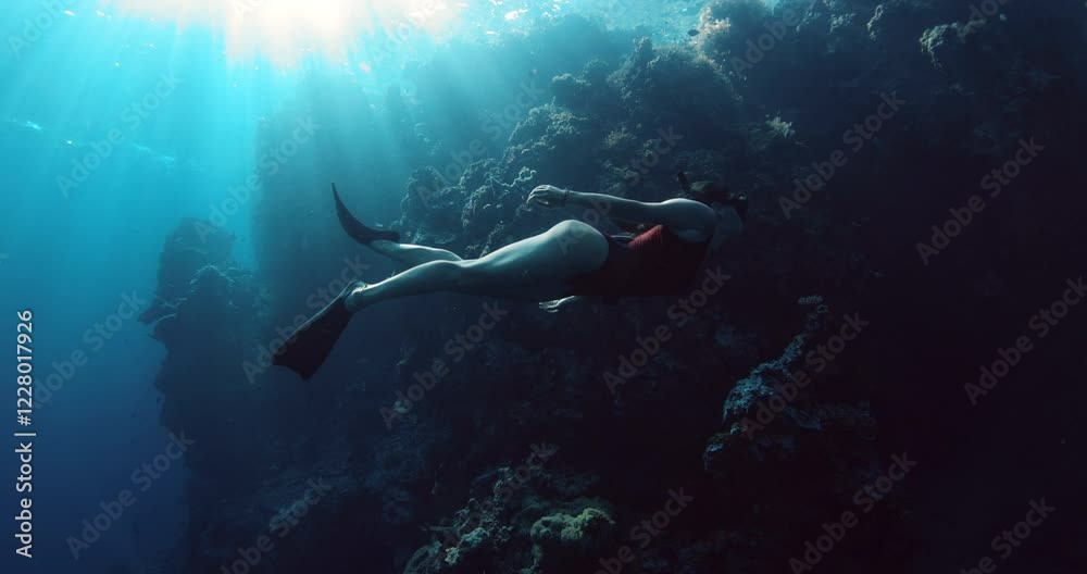 Freediver swims undersea in deep ocean with sun rays. Woman free diving in blue sea near rocks and coral reef Freediver swims undersea in deep ocean with sun rays. Woman free diving in blue sea near rocks and coral reef