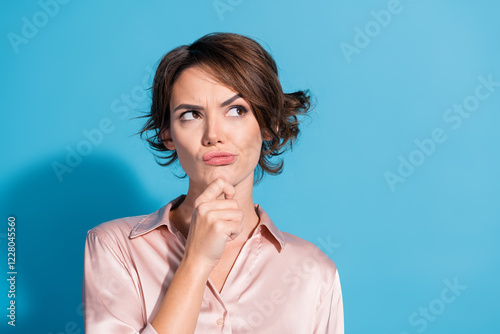 Young stylish woman in satin pink blouse pondering with funny face on blue background