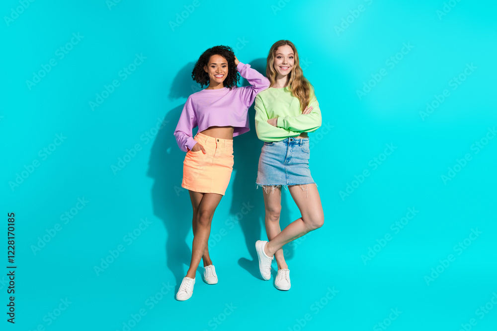 Obraz premium Two young women in trendy outfits posing against teal background, showcasing friendship, diversity, and modern fashion style.