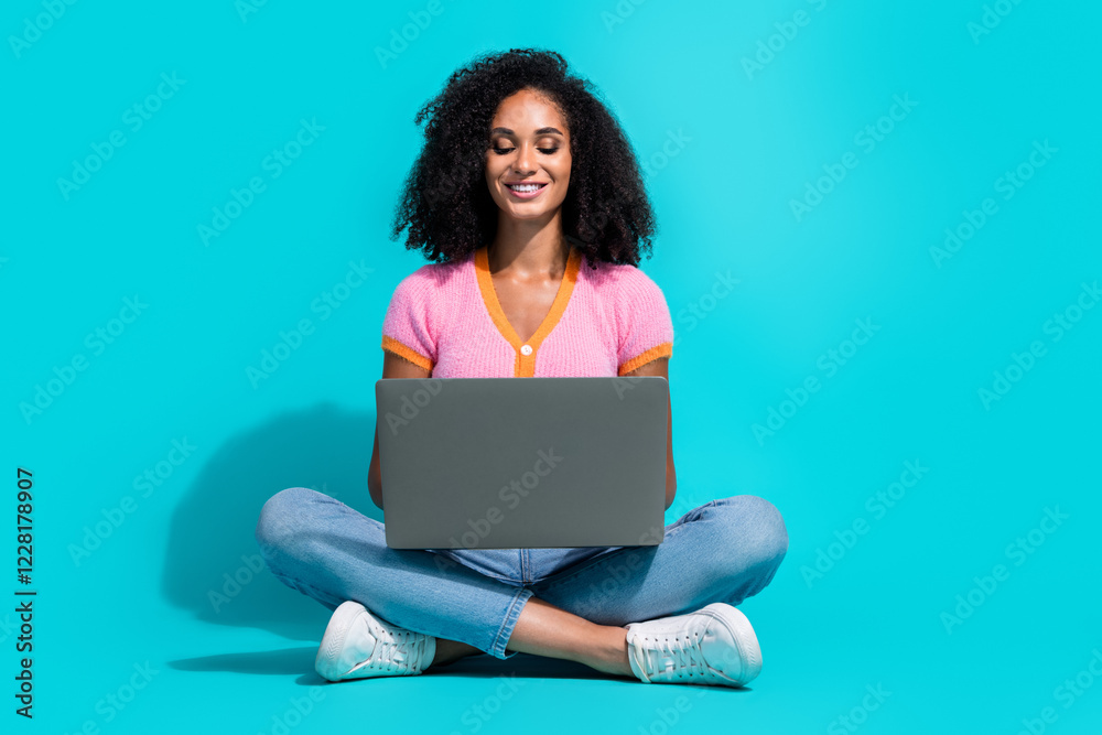 Naklejka premium Young woman sitting cross-legged, using laptop on turquoise background, wearing pink top and jeans, smiling and relaxed.