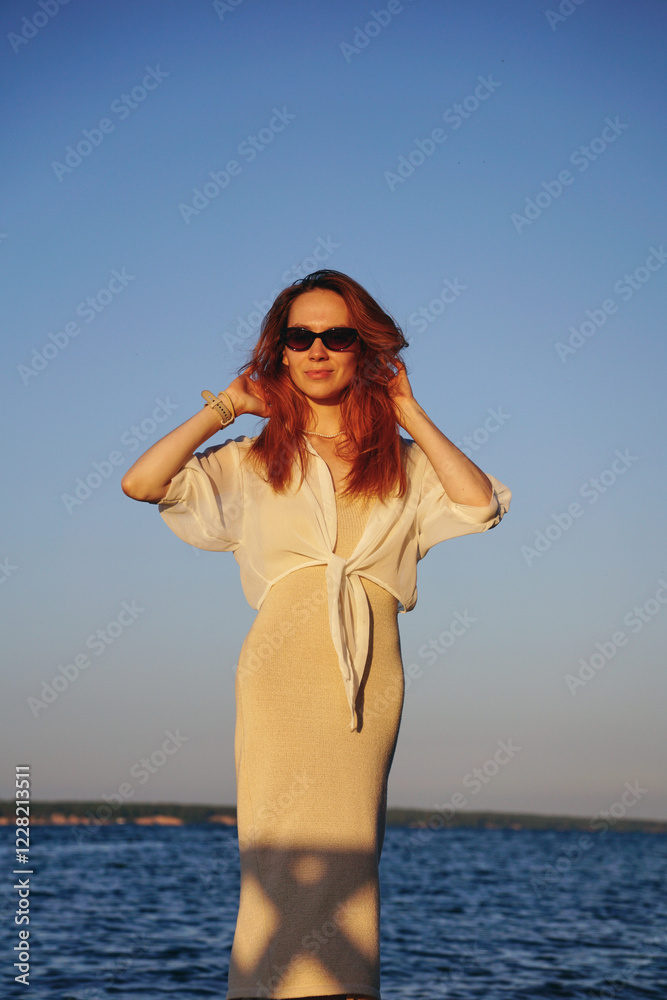 A young woman with long red hair in a tight dress stands on the beach by the river during sunset on a summer evening A young woman with long red hair in a tight dress stands on the beach by the river during sunset on a summer evening