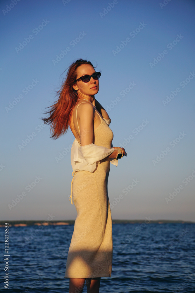A young woman with long red hair in a tight dress stands on the beach by the river during sunset on a summer evening A young woman with long red hair in a tight dress stands on the beach by the river during sunset on a summer evening