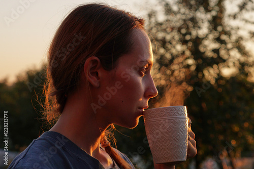 Obraz na plátne A young woman drinks hot tea from a steaming mug - A cozy summer evening at suns