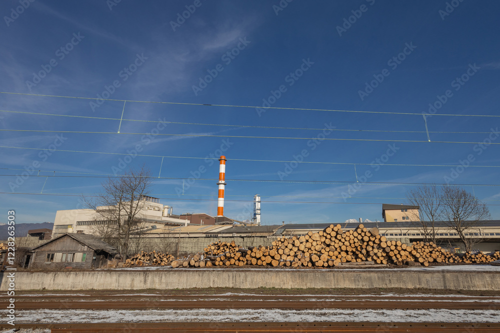 A pile of logs on the train station of Skofja Loka, Slovenia. Industrial facility in the background with visible red and white chimney.