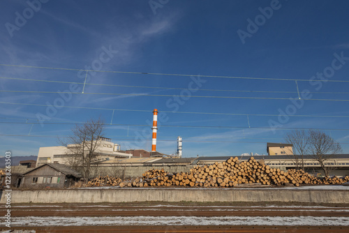 Obraz na płótnie A pile of logs on the train station of Skofja Loka, Slovenia. Industrial facility in the background with visible red and white chimney. w salonie