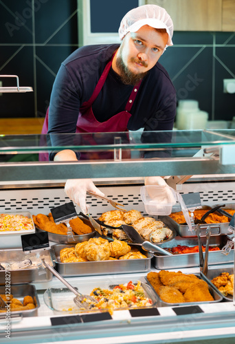 Фотография Polite young bearded salesman in apron and hairnet serving various ready-to-eat dishes from refrigerated deli counter in supermarket