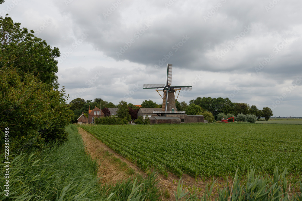Windmill stands tall against a cloudy sky in a rural landscape with lush green fields and winding path Windmill stands tall against a cloudy sky in a rural landscape with lush green fields and winding path
