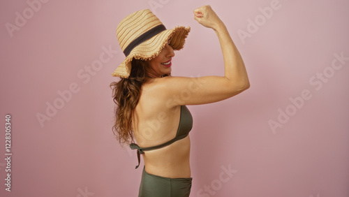 Woman flexing in swimsuit with sunhat and pink background showing confidence and strength