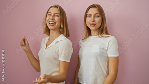 Two smiling blonde sisters stand together in front of a pink isolated wall, radiating happiness and familial love, both wearing casual white outfits, embracing their bond.