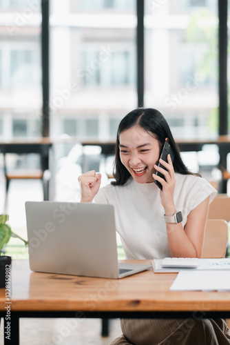 A joyful young woman celebrates success on a phone call while working on her laptop in a bright, modern office setting.