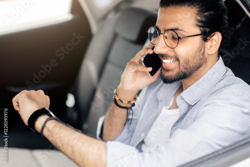 Happy young indian man in casual outfit sitting at car back seat, having phone conversation, checking time, going to airport by taxi, middle-eastern guy taking cab, copy space photo