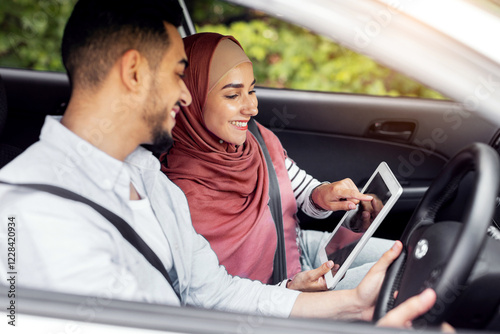 Cheerful millennial muslim female in hijab shows way on tablet with empty screen to husband at steering wheel in car, outdoor. Digital map, online application for travel, device and road to vacation photo