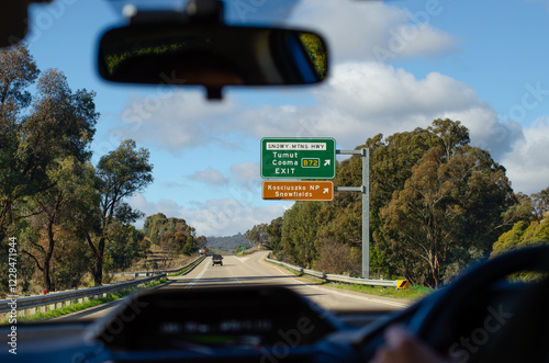 A driver's perspective view through the windshield of a car shows a highway road with directional signs for Tumut, Cooma, and Kosciuszko National Park snow fields on the roadside. NSW, Australia. photo