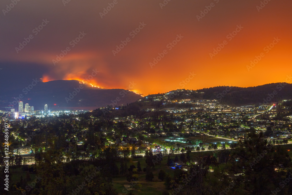 Obraz premium A wildfire burns on a mountainside near Kelowna illuminating the night sky with an orange glow while city lights shine below.