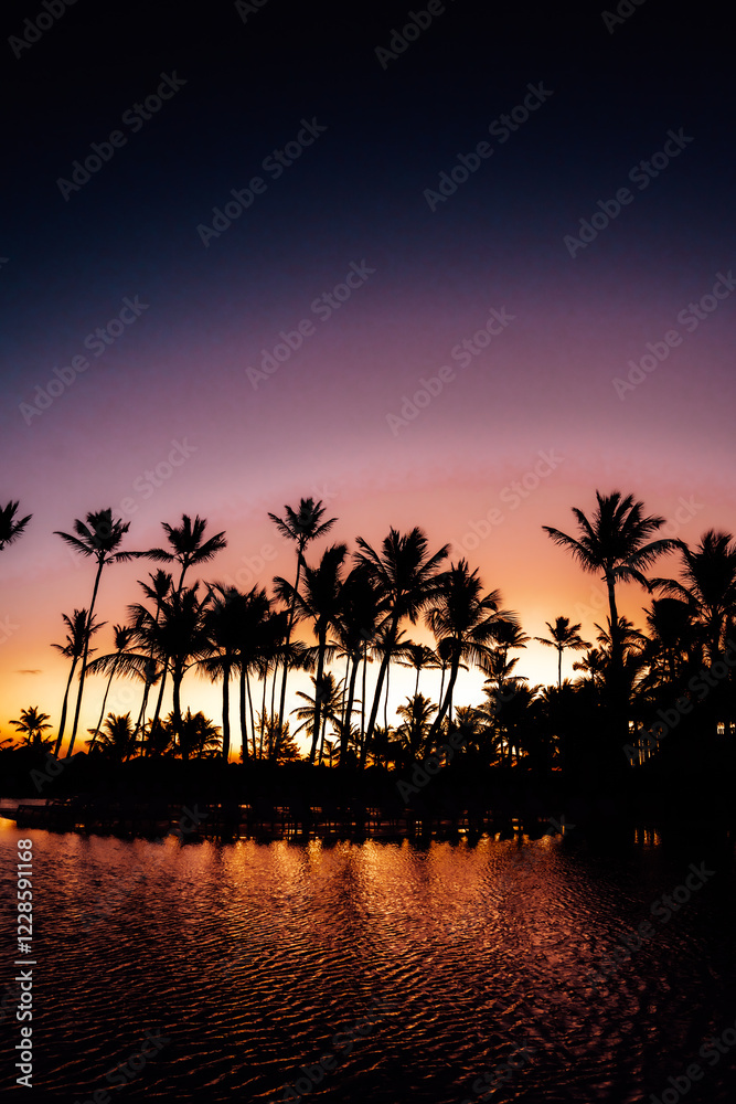 Obraz premium An early morning tropical sunrise with a background of tall palm trees and water ripples on a pool showing bright orange and yellow colors.