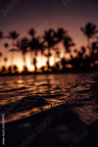 An early morning tropical sunrise with a background of tall palm trees and water ripples on a pool showing bright orange and yellow colors.