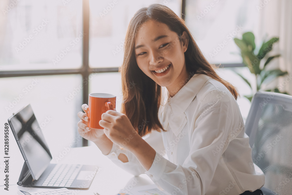 Young asian businesswoman sitting on an ergonomic chair and drinking coffee while working on a laptop computer in a modern office, she is smiling and looking at the screen Young asian businesswoman sitting on an ergonomic chair and drinking coffee while working on a laptop computer in a modern office, she is smiling and looking at the screen