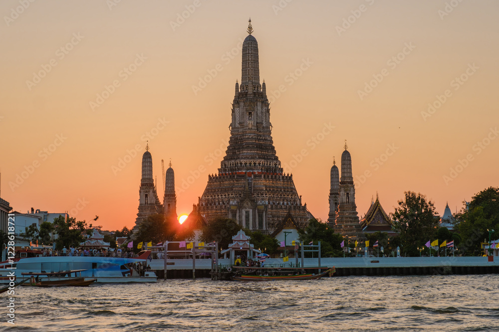 Fototapeta premium Golden rays of the setting sun illuminate the majestic Wat Arun temple, casting stunning reflections on the Chao Phraya River in Bangkok. The vibrant hues create a magical atmosphere.