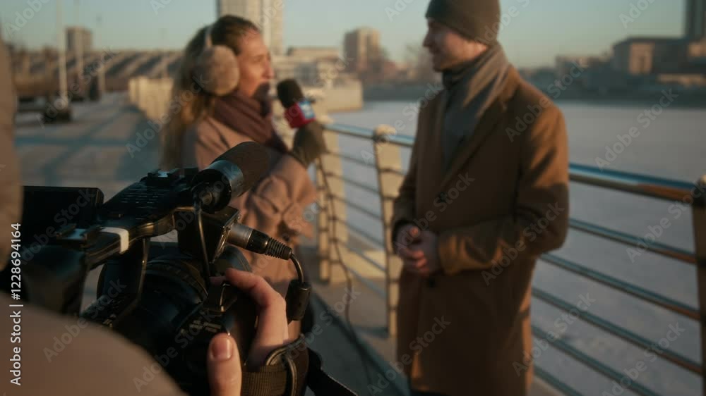 Over-shoulder shot of anonymous TV operator with camera filming young Caucasian female reporter talking to male local council head in smart winter coat while standing on river bridge Over-shoulder shot of anonymous TV operator with camera filming young Caucasian female reporter talking to male local council head in smart winter coat while standing on river bridge