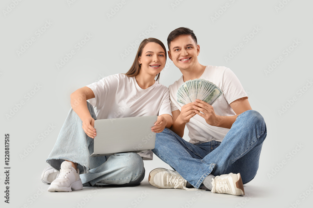 Young couple with money and laptop sitting on light background