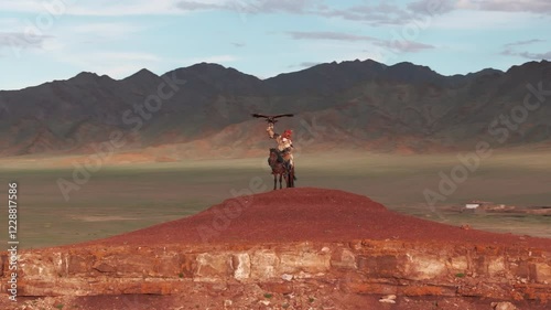 Aerial drone view of a traditional Mongolian eagle hunter on horseback, climbing a hill with the eagle in his arms, spectacular shot with the steppe in the background at sunset