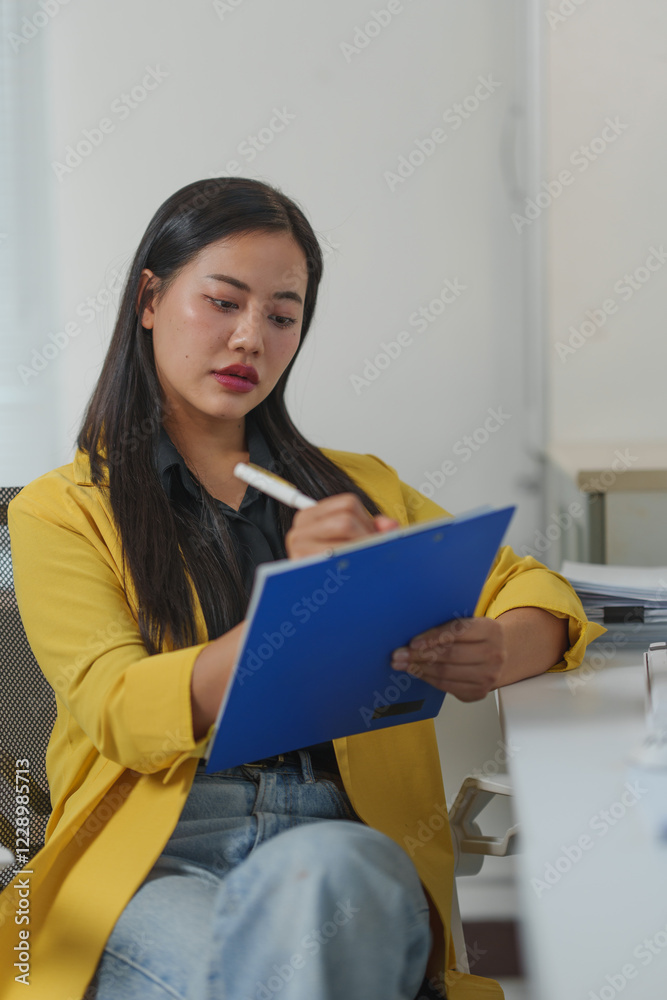 Young businesswoman in a yellow blazer writing on a clipboard while seated at a desk in a modern office. Concentrated on her task, she exemplifies professionalism and dedication Young businesswoman in a yellow blazer writing on a clipboard while seated at a desk in a modern office. Concentrated on her task, she exemplifies professionalism and dedication