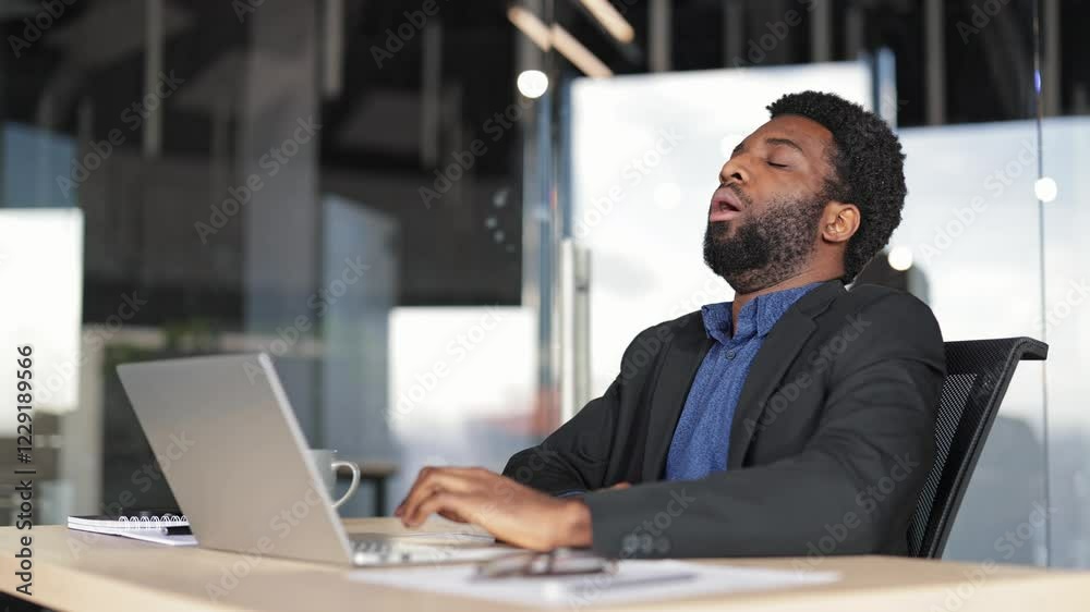 Tired businessman yawning at workplace in modern office. Exhausted African American male worker feeling sleepy, working on laptop computer. Tired businessman yawning at workplace in modern office. Exhausted African American male worker feeling sleepy, working on laptop computer.