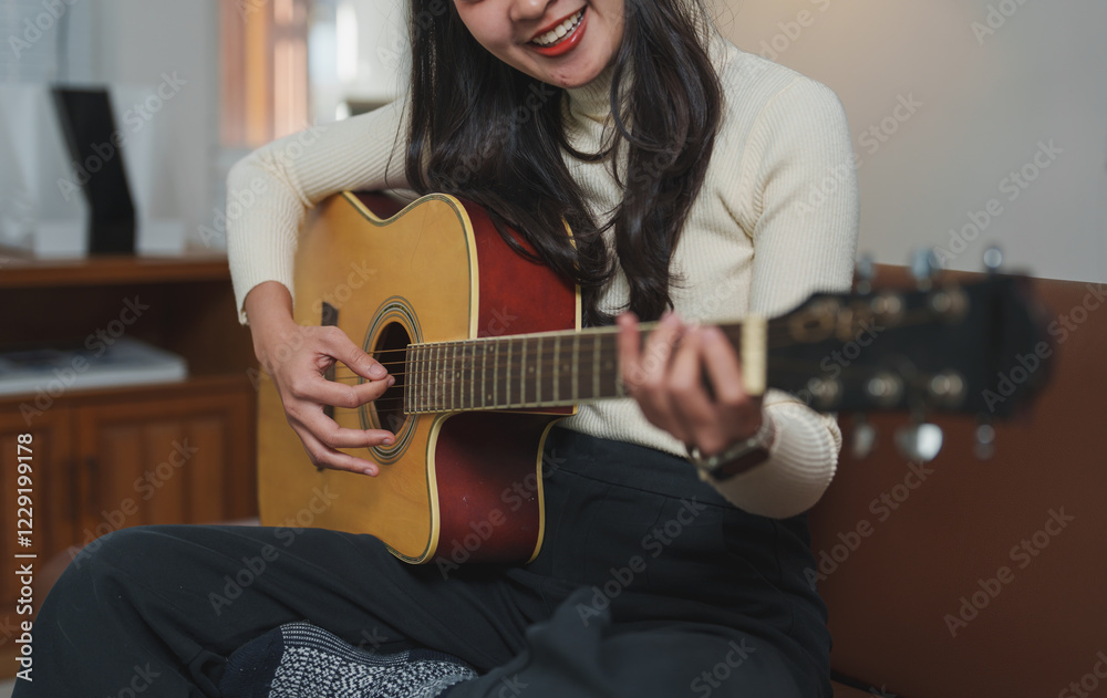 Obraz premium Woman sits comfortably on a couch, strumming an acoustic guitar with a smile. The cozy indoor setting enhances the warm, creative atmosphere, highlighting her passion for music