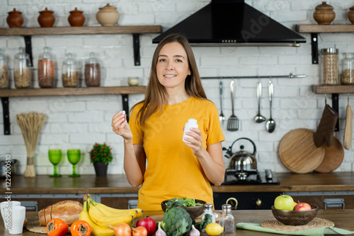 Vitamins, nutritional supplements, healthy lifestyle concept. Young woman eating breakfast and taking vitamin sitting at home in the kitchen, smiling friendly