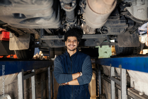 Portrait of diverse professional technician standing under chassis in garage trench with arms crossed and smiling at camera.