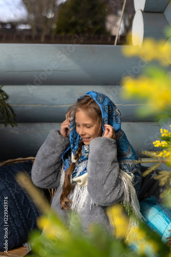 A beautiful Russian girl in a fur coat and headscarf sits on the porch of a wooden house on a sunny spring day.Family values,traditions and home comfort.The girl covers her hair with a beautiful scarf