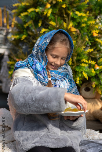 A beautiful Russian girl in a fur coat and a headscarf is sitting on the porch of a wooden house. Family values, traditions and home comfort.Maslenitsa holiday, winter send-off and spring meeting
