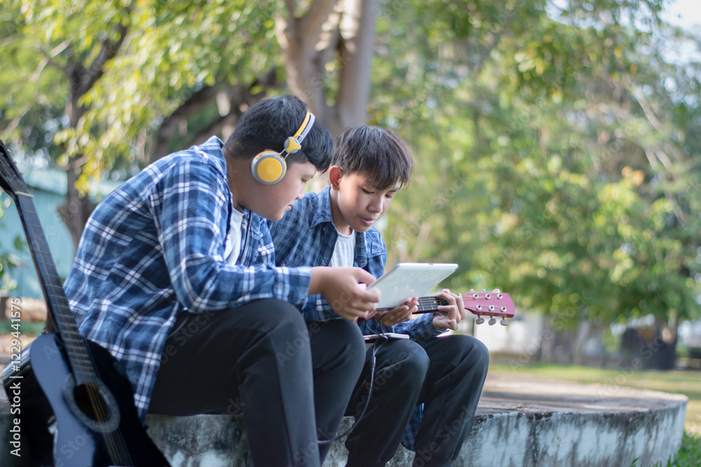 Fototapeta premium Asian teenaged boy spending his free times with his friend on weekend by practising and playing acoustic guitar or ukulele in the park together, recreational activity of teenagers concept.