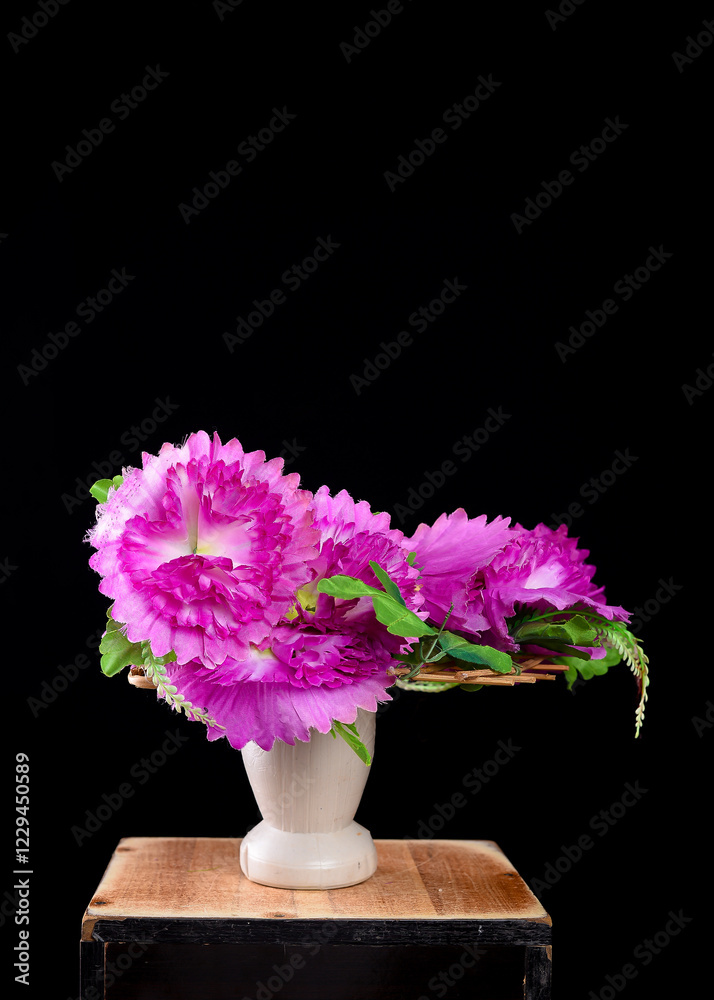 Obraz premium Bright pink flowers with ruffled petals arranged in a white vase, set against a black background on a wooden surface.